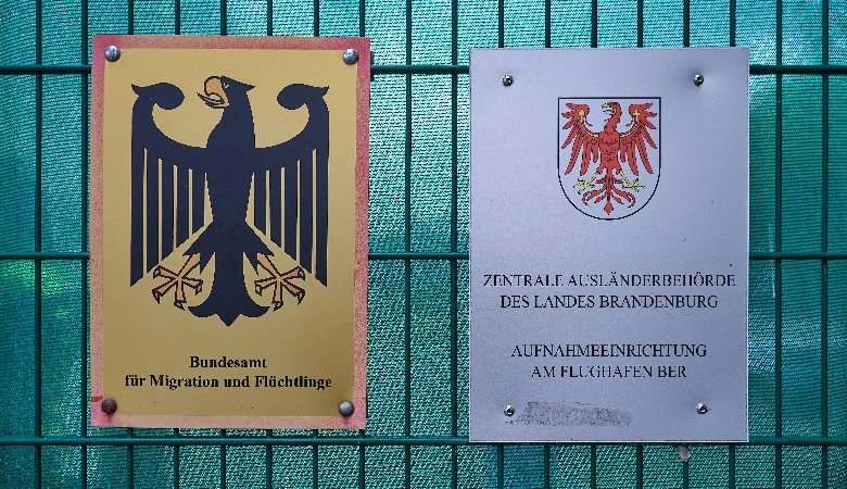 Schilder des Bundesamts für Migration und Flüchtlinge (BAMF) und der Zentralen Ausländerbehoerde des Landes Brandenburg (ZABH) am Zaun der neuen Außengrenzeinrichtung des Landes Brandenburg am BER-Flughafen.