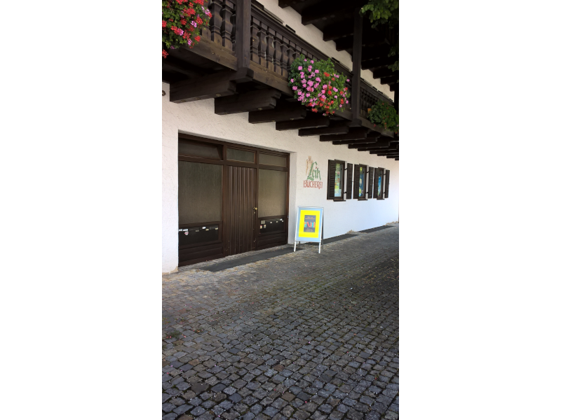 Eingang eines Gebäudes mit Holzbalkon und blühenden Blumen, Pflastersteinstraße, Schild mit Buchplakat vor der Tür.