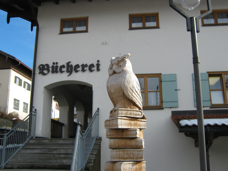 Holzeule auf einem Stapel Bücher vor einem Gebäude mit der Aufschrift 