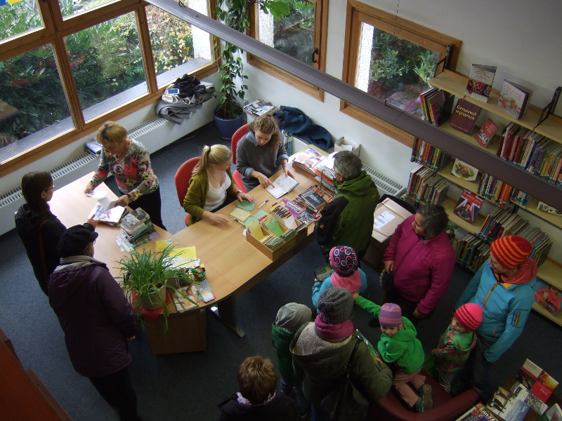 Menschen versammeln sich um einen Tisch in einer Bibliothek. Zwei Personen sitzen hinter dem Tisch und scheinen Bücher zu signieren oder zu verkaufen. Um den Tisch stehen mehrere Erwachsene und Kinder, einige tragen bunte Winterkleidung. Im Hintergrund si