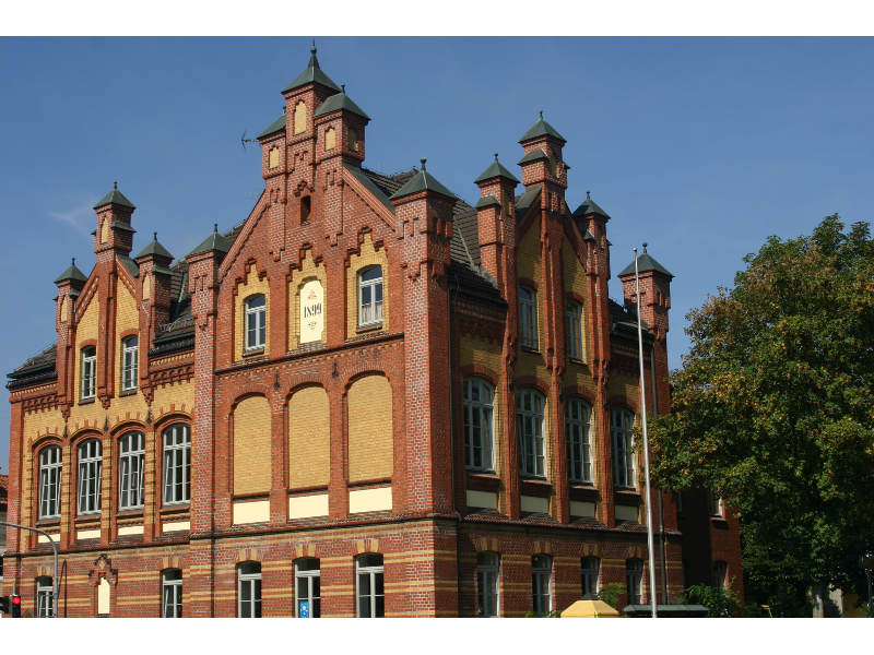Historisches rotes Backsteingebäude mit Türmen und Spitzdächern, umgeben von Bäumen, bei sonnigem Wetter.