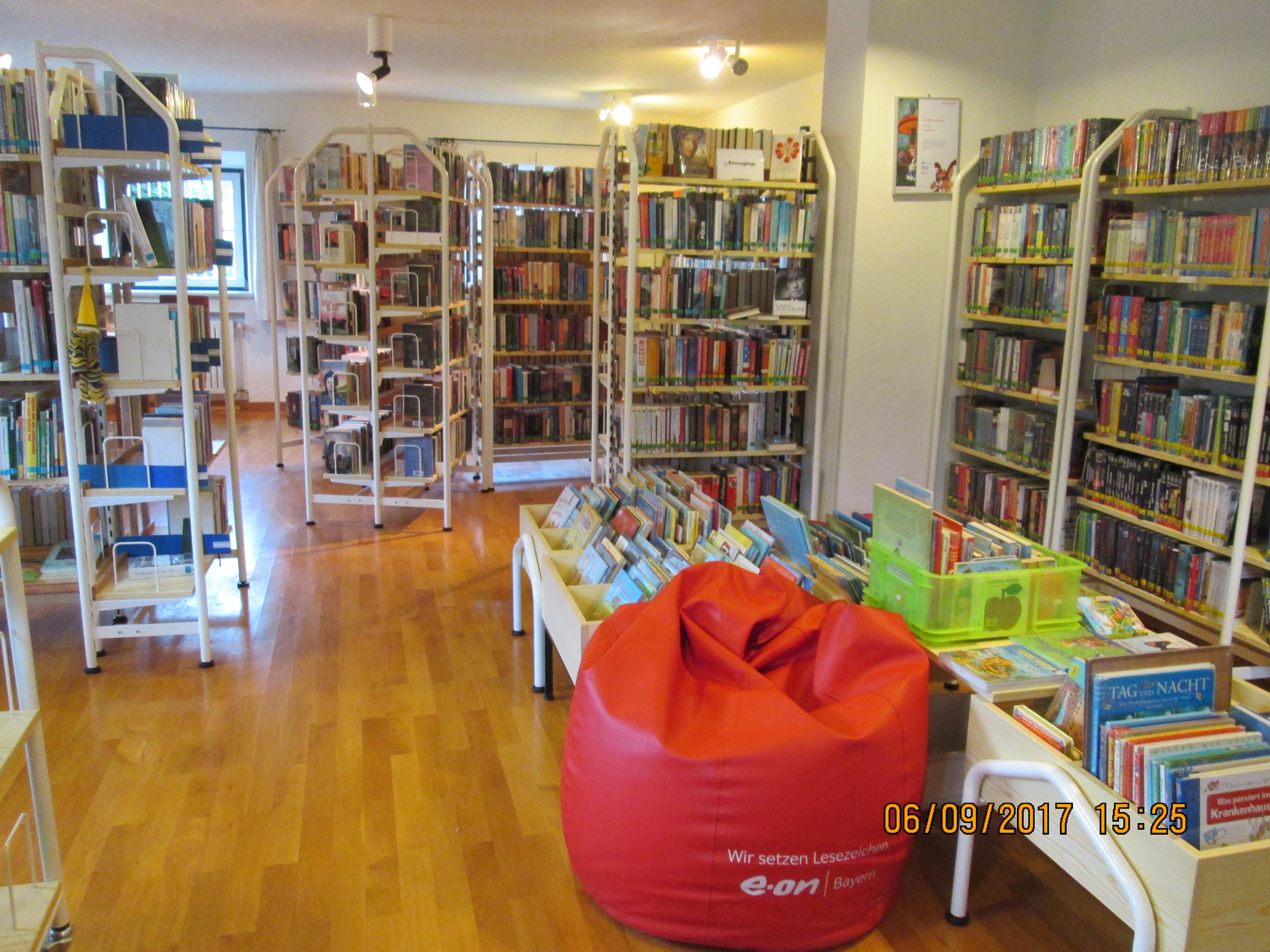 Innenansicht einer Bibliothek mit Holzfußboden. Regale voller Bücher säumen die Wände. In der Mitte steht ein roter Sitzsack mit der Aufschrift 