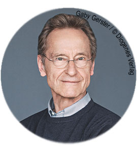 Ein älterer Mann mit Brille und grauem Haar, trägt einen dunkelblauen Pullover, lächelt leicht und blickt direkt in die Kamera.
