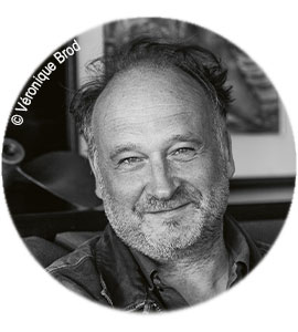 Schwarz-weiß Porträt eines lächelnden Mannes mit Bart, im Hintergrund unscharfe Kunstwerke.