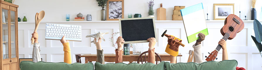 Personen hinter einem grünen Sofa halten verschiedene Gegenstände hoch: eine Computertastatur, einen Holzlöffel, ein Modellflugzeug, eine Tafel, ein Malbrett und eine Ukulele. Im Hintergrund sind Regale mit Dekorationen und Pflanzen zu sehen.